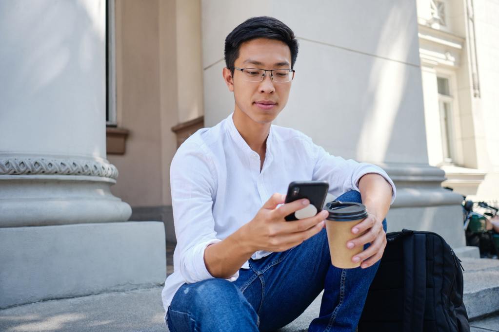 Asian student in eyeglasses with coffee to go thoughtfully using cellphone near university