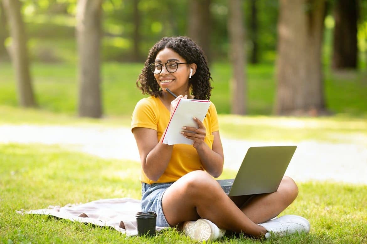 Online education. Happy black girl taking notes while watching web lesson on laptop at park